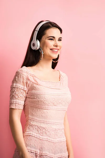 Alegre chica escuchando música en auriculares inalámbricos y mirando hacia otro lado en el fondo rosa - foto de stock