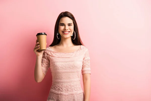 Cheerful girl smiling at camera while holding coffee to go on pink background — Stock Photo