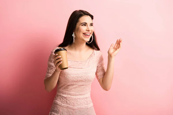 Glückliches Mädchen, das wegschaut und lächelt, während es Kaffee auf rosa Hintergrund hält — Stockfoto