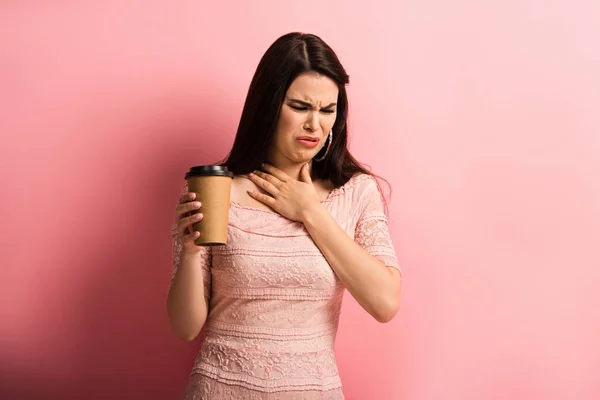 Недовольная девушка трогает грудь, держа кофе, чтобы пойти на розовый фон — стоковое фото