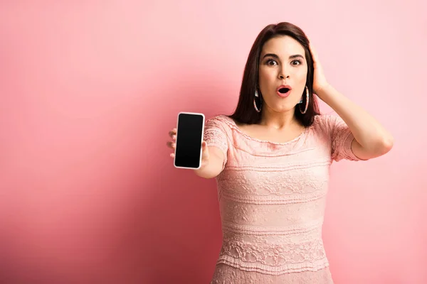 Chica impresionada tocando la cabeza mientras muestra el teléfono inteligente con pantalla en blanco sobre fondo rosa - foto de stock