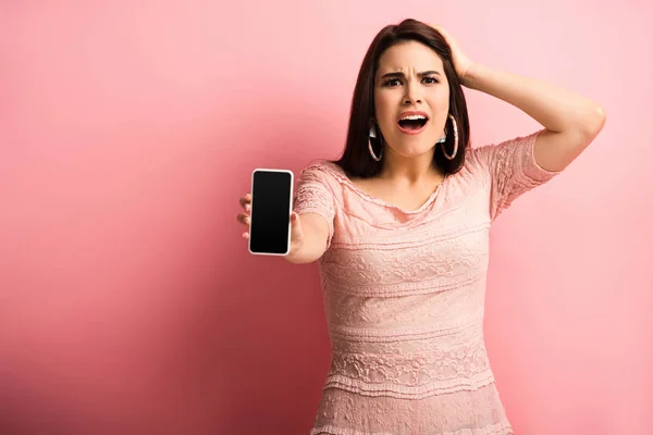 Chica conmocionada tocando la cabeza mientras muestra el teléfono inteligente con pantalla en blanco sobre fondo rosa - foto de stock