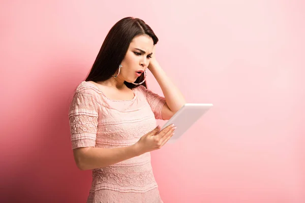 Chica conmocionada tocando el cabello mientras mira la tableta digital sobre fondo rosa - foto de stock