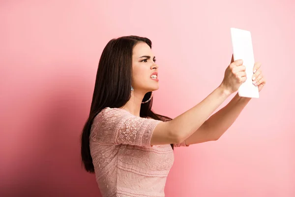 Angry girl looking at digital tablet on pink background — Stock Photo