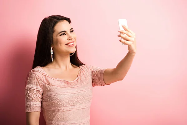 Щаслива дівчина посміхається, беручи селфі на смартфон на рожевому фоні — стокове фото