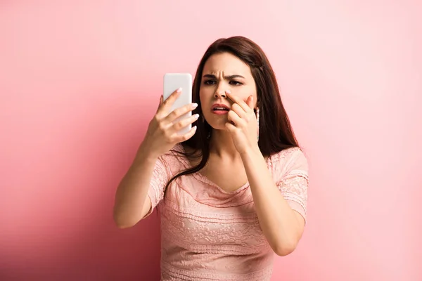 Недовольная девушка трогает нос, глядя на смартфон на розовом фоне — стоковое фото