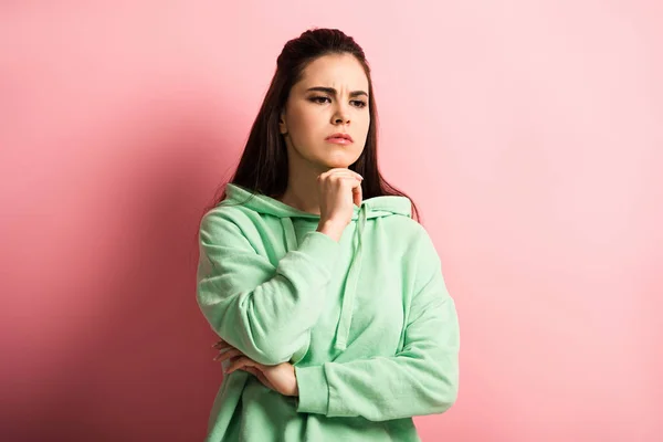 Upset, thoughtful girl holding hand near chin while standing on pink background — Stock Photo