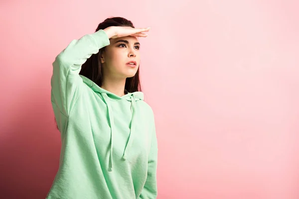 Focalizzata ragazza guardando lontano mentre si tiene la mano sotto gli occhi su sfondo rosa — Foto stock