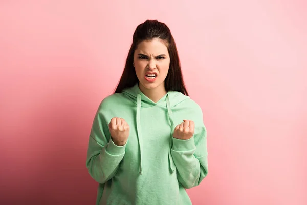 Сердитая девушка показывает сжатые кулаки, глядя на камеру на розовом фоне — стоковое фото