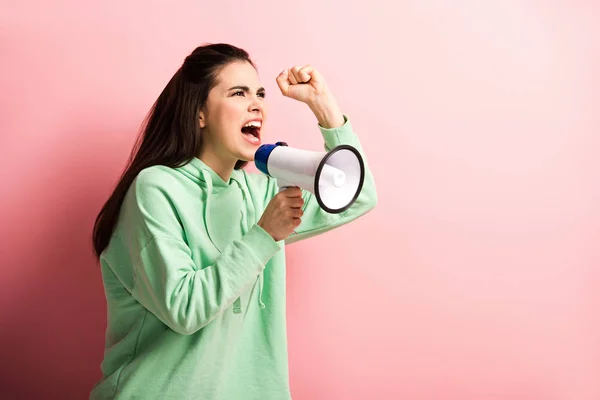 Menina zangada mostrando punho cerrado enquanto gritando em megafone no fundo rosa — Fotografia de Stock