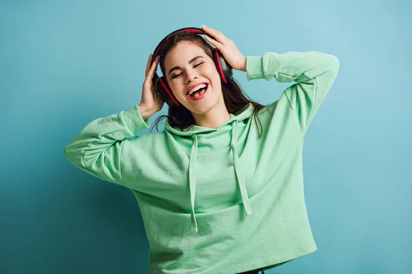 Chica excitada en auriculares inalámbricos cantando con los ojos cerrados sobre fondo azul - foto de stock