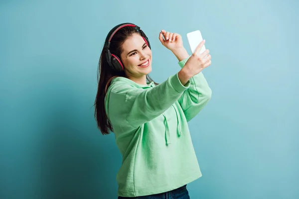 Chica feliz en auriculares inalámbricos tomando selfie en el teléfono inteligente sobre fondo azul - foto de stock