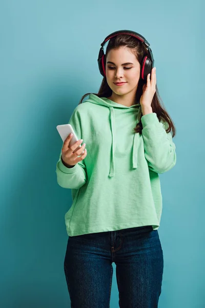 Menina sorridente em fones de ouvido sem fio olhando para smartphone no fundo azul — Fotografia de Stock