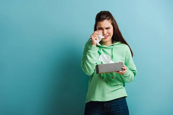 Chateado, chorando menina olhando para a câmera enquanto enxuga lágrimas com guardanapo de papel no fundo azul — Fotografia de Stock