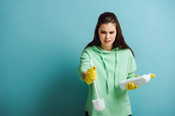 Ama de casa disgustado sosteniendo inodoro limpiador y émbolo sobre fondo azul — Stock Photo