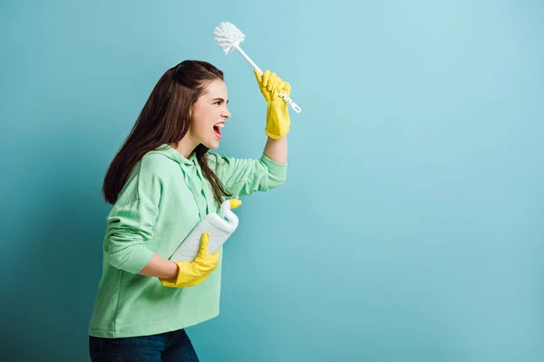 Seitenansicht einer wütenden Hausfrau, die einen Kolben in der erhobenen Hand hält und auf blauem Hintergrund schreit — Stockfoto