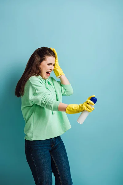 Descontento ama de casa mueca y tocando la cabeza mientras pulveriza ambientador sobre fondo azul - foto de stock