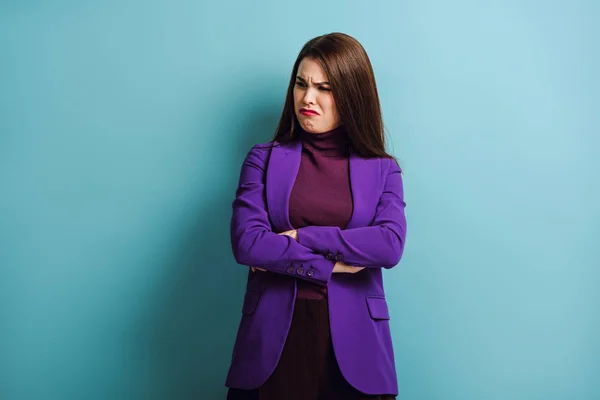 Sad young woman crying while standing with crossed arms on blue background — Stock Photo