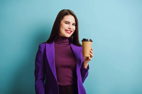 Menina alegre olhando para a câmera enquanto segurando café para ir no fundo azul — Fotografia de Stock