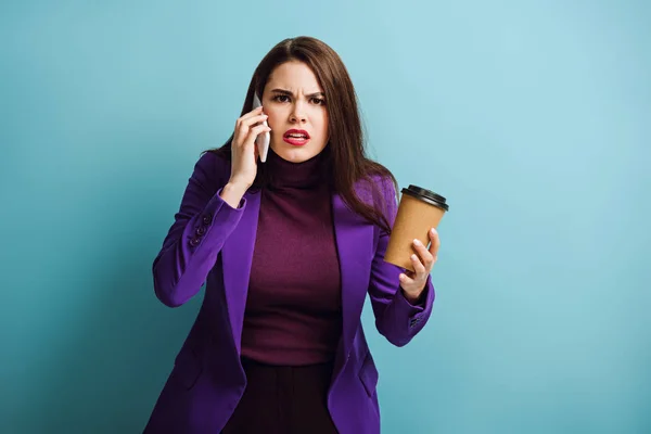 Сердитая девушка разговаривает по смартфону и держит кофе на синем фоне — стоковое фото