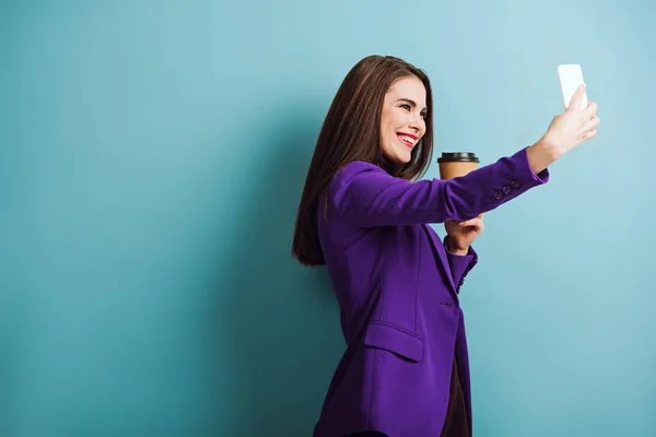 Весела дівчина бере селфі на смартфон, тримаючи каву, щоб йти на синьому фоні — стокове фото