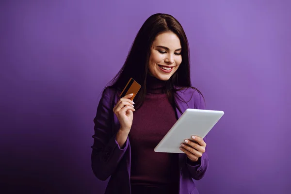 Mujer joven sonriente mirando la tableta digital mientras sostiene la tarjeta de crédito en el fondo púrpura - foto de stock