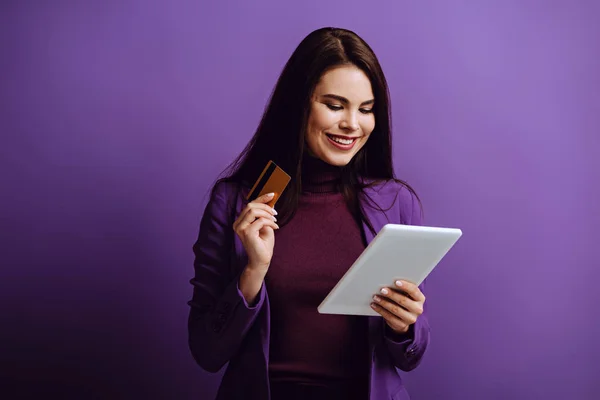 Mujer joven sonriente mirando la tableta digital mientras sostiene la tarjeta de crédito en el fondo púrpura — Stock Photo