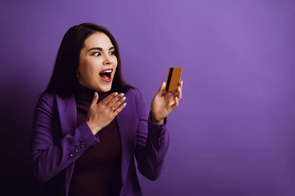 Шокована молода жінка тримає руку біля обличчя, дивлячись на кредитну картку на фіолетовому фоні — стокове фото