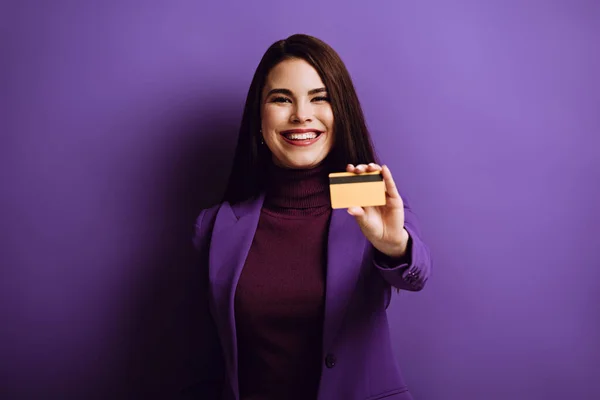 Glückliche junge Frau lächelt, während sie Kreditkarte auf lila Hintergrund zeigt — Stockfoto