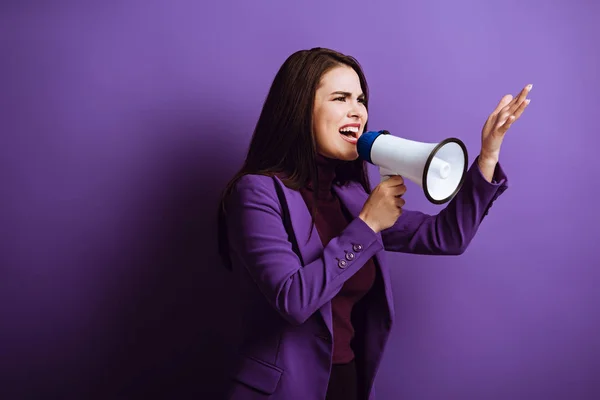 Angry young woman shouting in megaphone and showing indignation gesture on purple background — Stock Photo