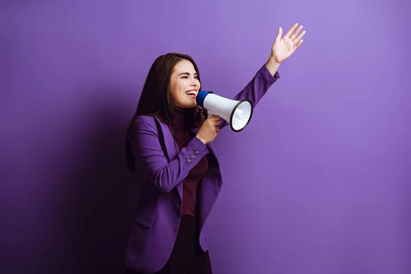 Excited young woman talking in megaphone while standing with raised hand on purple background — Stock Photo