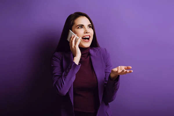 Impressed girl standing with open arm while talking on smartphone on purple background — Stock Photo