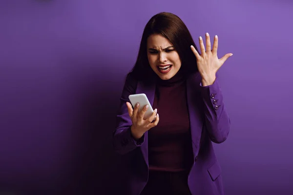 Angry young woman standing with raised hand while having video chat on purple background — Stock Photo