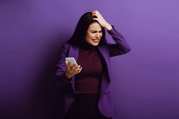Aufgebrachte junge Frau berührt Kopf, während sie Smartphone auf lila Hintergrund hält — Stockfoto