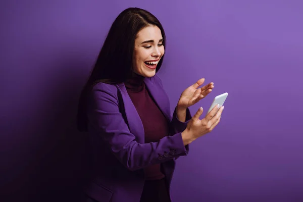 Chica emocionada de pie con el brazo abierto y riendo mientras tiene video chat sobre fondo púrpura - foto de stock