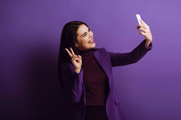 Happy young woman showing victory gesture while taking selfie on purple background — Stock Photo