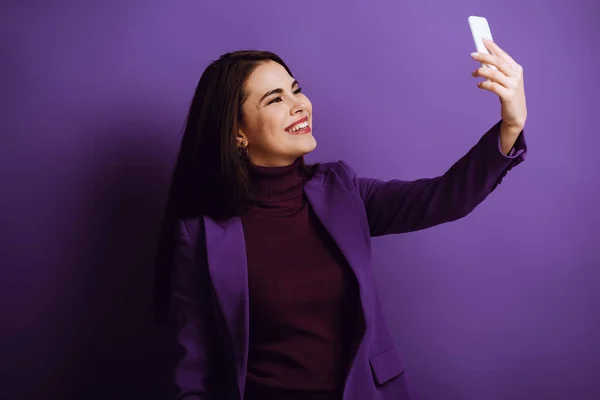 Весела стильна дівчина бере селфі на смартфон на фіолетовому фоні — стокове фото