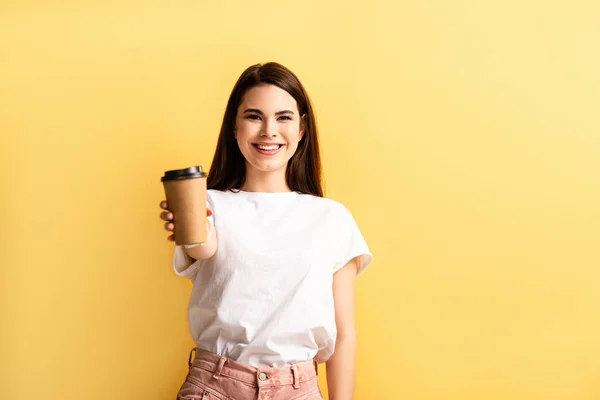 Glücklich attraktives Mädchen zeigt Coffee to go, während es in die Kamera lächelt, isoliert auf gelb — Stockfoto