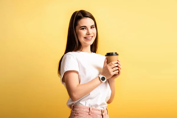 Весела дівчина тримає каву, щоб піти, дивлячись на камеру ізольовано на жовтому — стокове фото