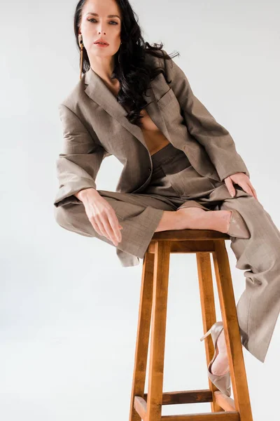 Mujer sexy y elegante en traje sentado en el taburete sobre fondo gris - foto de stock