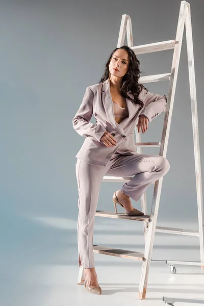Femme sexy et élégant en costume près de l'échelle en regardant la caméra sur fond gris — Photo de stock