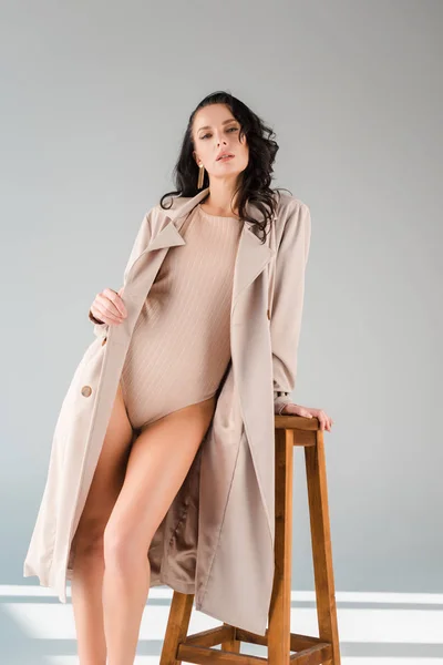 Atractiva mujer en traje de cuerpo y abrigo de pie cerca de taburete de madera sobre fondo gris - foto de stock