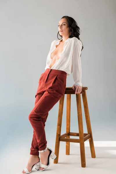 Atractiva y sexy mujer en camisa y pantalones de pie cerca de taburete sobre fondo gris - foto de stock