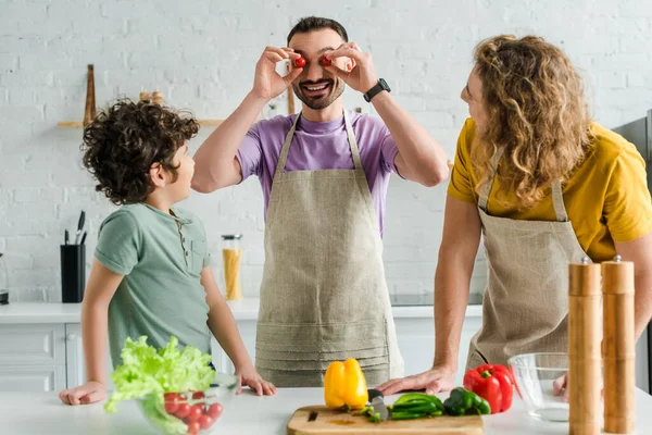 Feliz y barbudo homosexual hombre cubriendo los ojos con tomates cereza cerca de raza mixta hijo y pareja - foto de stock