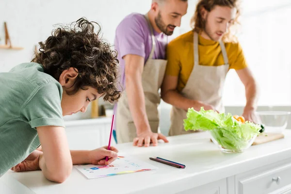 Enfoque selectivo del dibujo de niños de raza mixta con lápiz de color cerca de padres homosexuales - foto de stock