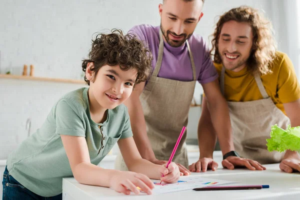 Selective focus of happy mixed race kid drawing picture near happy homosexual parents — Stock Photo