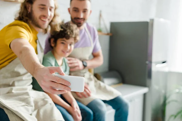 Enfoque selectivo de hombres homosexuales felices tomando selfie con lindo hijo de raza mixta - foto de stock