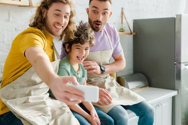 Selective focus of handsome homosexual men taking selfie with mixed race kid sticking out tongue — Stock Photo