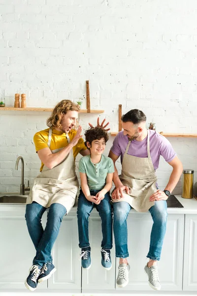 Happy homosexual parents sitting with mixed race son in kitchen — Stock Photo