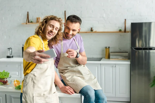 Enfoque selectivo de los hombres homosexuales sonrientes en delantales tomando selfie - foto de stock